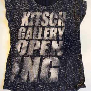 Svart t-shirt med tryck från New Style - Cool svart t-shirt från New Style med stort vitt texttryck 'KITSCH GALLERY OPENING' på framsidan. T-shirten har korta ärmar och ett unikt, nästan marmorerat mönster över hela tyget. Perfekt för dig som gillar statement-plagg och streetstyle.