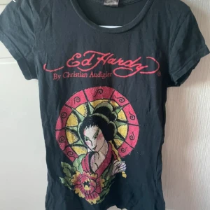 Svart Ed Hardy t-shirt  - Svart t-shirt från Ed Hardy med färgglatt tryck av en geisha och stora röda loggor fram och bak. T-shirten har korta ärmar med glittriga detaljer på motivet. Snygg och unik stil för dig som gillar statement-plagg. 