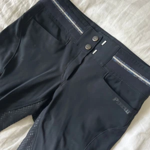 Svarta ridbyxor från Pikeur med silikon - Svarta ridbyxor från Pikeur med hög midja och silikongrepp längs hela benen. Byxorna har dubbla knappar i midjan, diskreta fickor och en snygg randig detalj i midjebandet. Stretchigt material som ger en tight och bekväm passform.