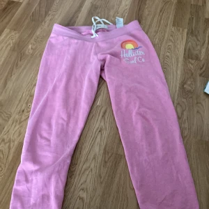 Rosa mjukisbyxor från Hollister  - Rosa mjukisbyxor från Hollister med regnbåge och med text som står 'Hollister Surf Co' på ena benet. Byxorna har ett justerbart snöre i midjan och är i mjukt bomullsmaterial. Perfekt som myskläder eller till och med idrotten!