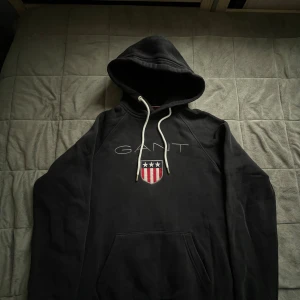 Svart hoodie från GANT med logga XS - Svart hoodie från GANT i storlek XS med broderad GANT-logga och sköld på bröstet. Tröjan har huva med vita snören och en stor magficka. 
