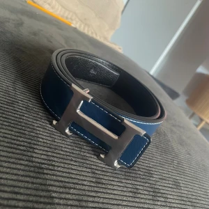 H Bälte - Snyggt blått skärp från Hmed ikoniskt H-spänne i silverfärgad metall. Bältet har vita kontrastsömmar och är gjort i skinn med svart insida. Perfekt accessoar för att lyfta din outfit.