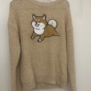 Beige stickad tröja med hundmotiv - Säljer en beige stickad tröja med ett stort, gulligt hundmotiv framtill. Tröjan har lång ärm och är gjord i ett grovstickat material som känns både mysigt och trendigt.