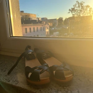 Svarta hasbeens i skinn, träbotten stl 36 - Snygga svarta sandaler med flätade remmar i skinn och justerbart spänne runt ankeln. Sulan är i ljust trä med gummidetalj under hälen för extra grepp. Perfekta för varma dagar och ger en stilren vibe till din outfit.