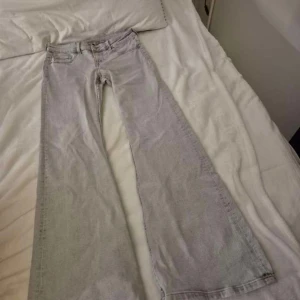 Ljusgråa Low waist bootcut jeans - Säljer ett par ljusgråa jeans med bootcut-modell och klassisk femficksdesign. Jeansen har bälteshällor och dragkedja med knapp framtill. Perfekta för dig som gillar en retroinspirerad look med breda ben och snygg passform.