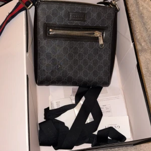 Gucci axelväska med GG-mönster - Snygg svart axelväska från Gucci med klassiskt GG-mönster och silverfärgad dragkedja framtill. Väskan har en bred axelrem i marinblått och rött, samt detaljer i skinn. Perfekt för dig som vill ha en stilren och ikonisk accessoar…. Måste gå till skräddaren klippa bort lite av bandet inget som är wow stort kostar 100kr eller under hundra spänn äkta kvitto finns till äkta allt ingår till väskan plus som sagt den måste fixas det syns på bilderna rakt o tydligt så läs innan du köper 