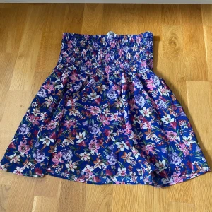 Blommig kortkjol från ONLY - Säljer en färgglad blommig kortkjol från ONLY med smockad midja. Kjolen har en blå bas med lila, rosa och vita blommor över hela tyget. Den är luftig och har en skön passform som sitter snyggt i midjan. Perfekt för dig som gillar mönster och färg!