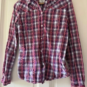 Rutig skjorta från L.O.G.G. H&M - Snygg rutig skjorta från L.O.G.G. by H&M i storlek 40. Skjortan har ett klassiskt rutmönster i rött, vitt och blått, med långa ärmar och knappar framtill. Tillverkad i mjuk bomull, perfekt för en chill och avslappnad stil.