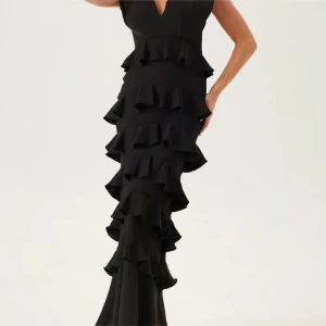 Svart långklänning med volanger - Supersnygg svart långklänning med djup v-ringning och flera lager volanger längs hela kjolen. Klänningen har ärmlös design och markerad midja, vilket ger en elegant och dramatisk siluett. Perfekt för dig som vill sticka ut med en unik look.