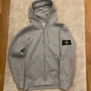 Grå hoodie från Stone Island - Säljer en grå hoodie från Stone Island med klassisk logotyp-patch på vänster ärm. Tröjan har huva med snörning, dragkedja framtill och två stora fickor. Perfekt för dig som gillar streetwear och vill ha något stilrent och bekvämt. Köptes från en kompis, därför osäkerhet av äktheten.