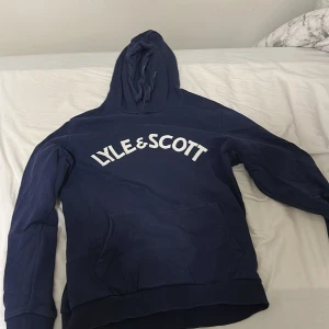 Marinblå hoodie från Lyle & Scott - Snygg marinblå hoodie från Lyle & Scott med stor broderad logga i vitt över bröstet. Tröjan har huva med dragsko och en stor magficka framtill. Perfekt för dig som gillar en avslappnad och sportig stil.
