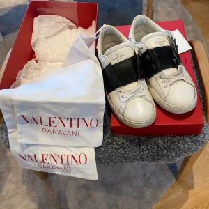 Valentino Garavani Open sneakers 41,5 äkta - Säljer ett par Valentino Garavani Open sneakers i vitt skinn med bred svart läderdetalj över vristen. Klassisk rund tå, platt sula och snörning. Kommer med originalkartong och dustbags. Perfekta för dig som vill ha en lyxig och stilren sneaker. köpta på Cettire, äkta