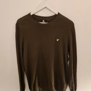 Brun stickad tröja från Lyle & Scott - Säljer en brun stickad tröja från Lyle & Scott med rund hals och klassisk gul logga på bröstet. Tröjan har lång ärm och är tillverkad i mjukt material som passar perfekt till lager-på-lager. Enkel och stilren design som funkar året runt.