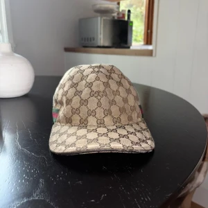 Gucci beige keps med GG-mönster - Säljer en beige keps från Gucci med det klassiska GG-monogrammet över hela kepsen. Kepsen har gröna och röda ränder på sidan och justerbar rem i svart skinn baktill. Materialet är bomull och skinn, perfekt för dig som vill ha en lyxig och snygg accessoar. Finns lite fläckar men går att tvätta bort
