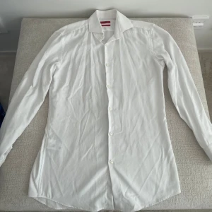 Vit slim fit skjorta från Hugo Boss - Vit skjorta från Hugo Boss i slim fit-modell med klassisk krage och knappar framtill. Skjortan är tillverkad i ett lätt bomullstyg som är easy iron, vilket gör den enkel att hålla snygg. Perfekt för dig som gillar stilrena och enkla plagg.