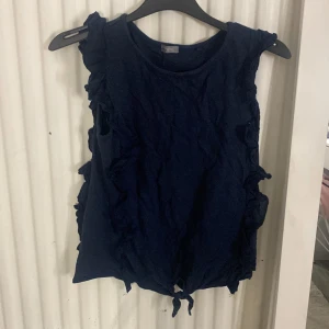 Mörkblå ärmlös topp med volanger - Supersöt mörkblå topp från Next med volangdetaljer längs sidorna och knytning längst ner. Toppen är ärmlös och har en rund halsringning. Perfekt för dig som gillar en lekfull och trendig stil.