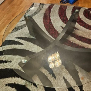Glass bord - Bilderna visar ett runt soffbord med glasskiva och svart korsad träbas. Bordet har en modern design och ser ut att vara i gott skick utan synliga repor eller skador på glaset eller benen. Perfekt för vardagsrummet och ger ett stilrent intryck.