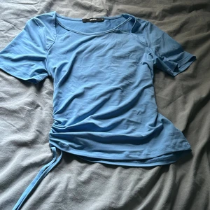 Blå topp med snörning  - Säljer en ljusblå topp från BikBok med kort ärm och cut out-detalj vid axlarna. Toppen har snörning och rynk på ena sidan som ger en snygg och trendig look💕