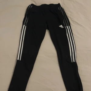 Svarta Adidas träningsbyxor med ränder - Svarta träningsbyxor från Adidas med klassiska vita ränder längs sidorna och Adidas-logga på låret. Byxorna har smal passform, resår i midjan och är gjorda i ett mjukt syntetmaterial som andas. Perfekta för träning eller chill.