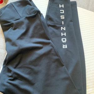 Svarta leggings från Rohnisch - Svarta leggings från Rohnisch med vit logga längs benet. De har en hög midja och är tillverkade i ett stretchigt material som passar perfekt för träning. Snygg och enkel design som är lätt att matcha med andra träningskläder.
