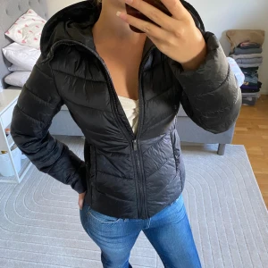 Svart dunjacka med fuskpäls från  - Snygg svart dunjacka från med avtagbar huva och fuskpäls. Jackan har quiltad design, dragkedja framtill och ribbade partier i sidorna för en schysst passform. Perfekt för kyliga dagar och riktigt stilren look.