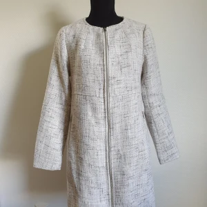 Beige kappa med dragkedja strl 34 - Stilren beige kappa i rak modell med diskret mönster och hel dragkedja framtill. Jackan har rund halsringning, långa ärmar och två fickor med dragkedja. Perfekt för dig som gillar minimalistisk och clean stil. Kappan är endast använd 1 gång. 