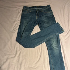 Nudie Jeans blå skinny jeans W27 L34 - Säljer ett par klassiska blå skinny jeans från Nudie Jeans med snygga slitningar och kontrastsömmar. Jeansen har den ikoniska Nudie-bågen på bakfickorna och är tillverkade i mjukt denim. Perfekt passform med smal siluett och normal midja.