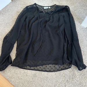 Svart blus med knyt från VILA - Säljer en svart transparent blus från VILA med diskret prickigt mönster och knytband vid halsen. 