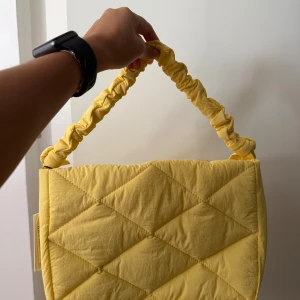 Gul quiltad handväska från Beck Söndergaard - Quiltad handväska i en härlig ljusgul färg från Beck Söndergaard. Väskan har ett vadderat, rutmönstrat tyg och ett scrunchie-inspirerat handtag. Stängs med dragkedja och har en rymlig insida med innerfack. Perfekt för dig som vill sticka ut med en färgglad accessoar.