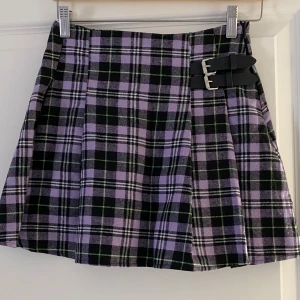 Lila rutig kortkjol från SHEIN - Superfin kortkjol från SHEIN i lila, svart och grönt rutigt mönster. Kjolen har två snygga spännen i midjan och är lätt utställd med veck. Perfekt för dig som gillar en cool och trendig stil.