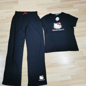 Svarta Hello Kitty 2 delar pyjamas  - Svarta pyjamas från Hello Kitty i 100% bomull med röd snörning i midjan och tryckt Hello Kitty-motiv vid benslutet. Byxorna har rak passform och är perfekta för chill dagar hemma.