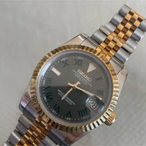 Seiko Datejust klocka i guld och silver - Snygg Seiko Datejust klocka med svart urtavla och romerska siffror. Armbandet är tvåfärgat i guld och silver, boetten har en räfflad guldkant. Klockan har datumvisning och klassisk design som sticker ut.