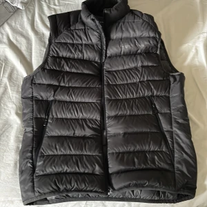 Svart dunväst från Sail Racing - Snygg svart dunväst från Sail Racing med quiltad design och hög krage. Västen har två dragkedjeförsedda fickor på sidorna och diskret logga på bröstet med en ytligare innerficka. Perfekt för lager-på-lager och håller dig varm under kyliga dagar. Pris går att förhandla 