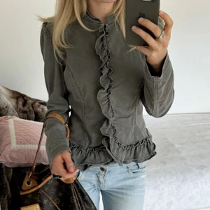 Khaki volangjacka i jeans - Säljer en grå jacka i bomull med volangdetaljer längs knäppningen och nederkanten. Jackan har lång ärm, figurnära passform och en cool, lite militärinspirerad look. 