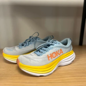 Hoka One One Bondi 8 ljusblå - Säljer ett par Hoka One One Bondi 8 i storlek 38. Skorna är ljusblå med gula och vita detaljer samt orange Hoka-logga. Ovandelen är i mesh för bra andningsförmåga och sulan är extra tjock för maxad komfort. Perfekta för dig som gillar snygga och bekväma träningsskor. Använda ett fåtal gånger inomhus. Säljer pga att dom är för små i storleken för mig. 