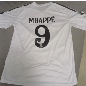 Real Madrid Mbappé fotbollströja - Snygg vit Real Madrid fotbollströja från Adidas med Mbappé och nummer 9 på ryggen. Tröjan har svarta detaljer, klubbmärke på bröstet och sponsortryck 'Emirates Fly Better'. Kortärmad modell i lätt funktionsmaterial, perfekt för match eller träning.