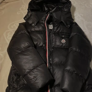 Svart Moncler pufferjacka med huva - Säljer en svart pufferjacka från Moncler med glansig finish, huva och klassisk logga på bröstet. Jackan har dragkedja med rödvit detalj, en bröstficka med knapp och vadderad design som håller dig varm. Perfekt för kalla dagar och riktigt snygg streetstil.
