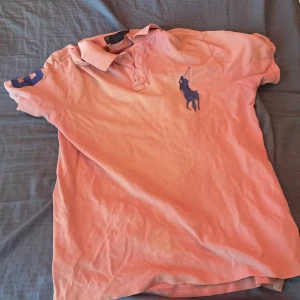 Rosa pikétröja från Polo Ralph Lauren - Säljer en rosa pikétröja från Polo Ralph Lauren med klassisk krage, korta ärmar och stor marinblå logga på bröstet. Tröjan är i 100% bomull och har en siffra broderad på ena ärmen. Perfekt för en chill och sportig look.