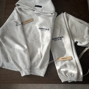 Grå Essentials Fear of God tracksuit  - Snygg ljusgrå hoodie från Essentials Fear of God med svart logga på bröstet. Klassisk känguruficka, huva och ribbade muddar. Perfekt för en clean och avslappnad streetstyle. Mjuk bomullsblandning som känns skön mot huden.
