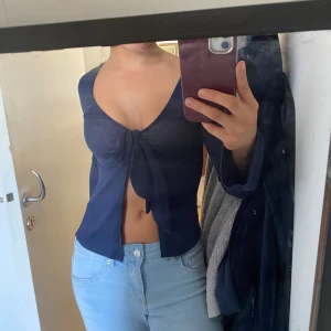 Mörkblå croppad knytblus - Säljer en mörkblå croppad blus med långa ärmar och djup v-ringning. Toppen har en snygg knytning framtill som ger en trendig look. Perfekt att styla med jeans eller kjol för en cool outfit. From ginatricot