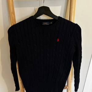 Mörkblå kabelstickad tröja Polo Ralph Lauren - Kabelstickad mörkblå tröja från Polo Ralph Lauren med klassisk röd broderad logga på bröstet. Tröjan har rund halsringning, lång ärm och ribbade muddar. Perfekt för lager-på-lager och snygg till jeans eller kjol.