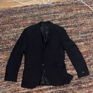 Svart kavaj från Jack & Jones Premium - Säljer en stilren svart kavaj från Jack & Jones Premium. Klassisk modell med två fickor fram, en bröstficka och knäppning med en knapp. Fodrad insida och fyra dekorativa knappar vid ärmslutet. Perfekt för dig som vill ha en clean och tidlös look.