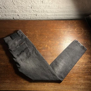 Svarta jeans från Nudie Jeans - (W30 L32) Snygga svarta jeans från Nudie Jeans med klassisk femficksdesign och diskreta sömmar på bakfickan. Jeansen har råa kanter vid benslut och är tillverkade i mjukt denimtyg. Perfekta för dig som gillar en stilren och avslappnad look.