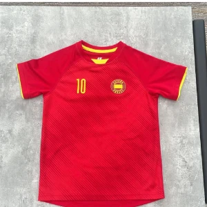 Röd Spanien fotbollströja H&M barn - Röd fotbollströja från H&M Sport med gula detaljer, nummer 10 och texten 'España' på ryggen. Tröjan har korta ärmar, rund hals med gul kant och subtila diagonala linjer i tyget. Perfekt för unga fotbollsfans som vill sticka ut på planen.
