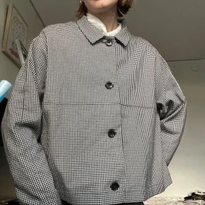 Svartvit rutig overshirt med knappar - Säljer en svartvit rutig overshirt med klassisk krage och stora svarta knappar framtill. Jackan har en boxig och loose passform, långärmade ärmar och är tillverkad i ett mjukt bomullstyg. Perfekt för lager-på-lager och ger en chill vibe till din outfit.