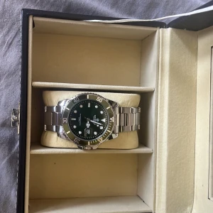 KOMPS svart och silver armbandsur - Stilren klocka från KOMPS med grön urtavla och silverfärgat armband i metall. Rund boett med tydliga siffror och datumvisning. Perfekt accessoar för dig som gillar klassisk design med modern touch. Snygg att bära både till skolan och på stan.