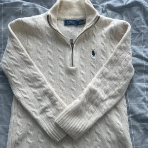 Vit kabelstickad tröja Polo Ralph Lauren - Vit kabelstickad tröja från Polo Ralph Lauren i ull och kashmir. Tröjan har half zip, hög krage och klassisk blå broderad logga på bröstet. Perfekt för dig som gillar en stilren och lyxig look med mjukt material och snygga detaljer.