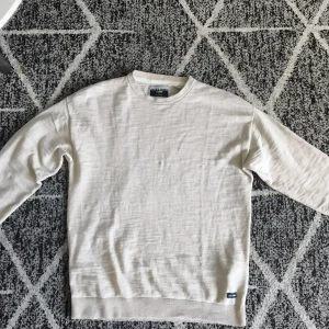 Beige tröja från Jack & Jones Core - En stilren beige tröja från Jack & Jones Core med rund halsringning och diskret logga nertill. Tröjan är långärmad och har ribbade muddar vid ärmslut och nederkant. 