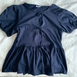 Mörkblå blus med knyt och volang - Supersöt mörkblå blus med puffiga korta ärmar, knyt framtill och volang nertill. Blusen har rund halsringning och är gjord i mjuk bomull, perfekt för en chill och trendig look.