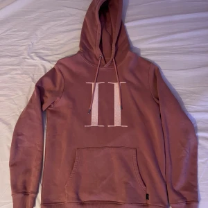 Rosa hoodie från Les Deux med tryck - Säljer en snygg rosa hoodie från Les Deux med stort vitt II-tryck på bröstet. Tröjan har huva med snörning, magficka och långa ärmar. Perfekt för dig som gillar stilrena och bekväma plagg med en modern touch.